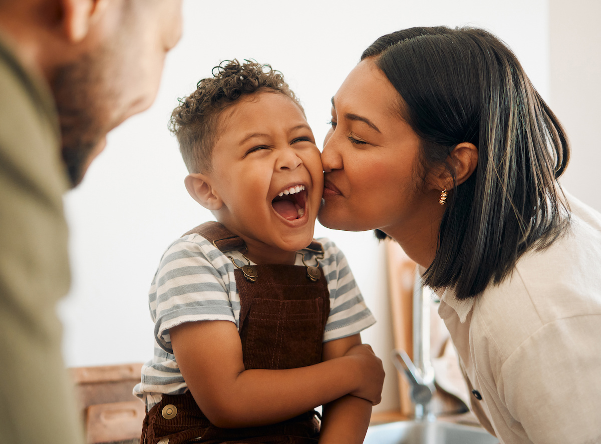 Happy boy getting a kiss by caring mother, bonding and laughing during family time at home. Young parents sharing a sweet moment of parenthood with their playful child, relaxing and carefree together