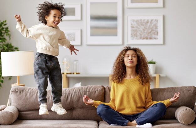 Young african woman mother in lotus pose meditating while child playing on background in living room